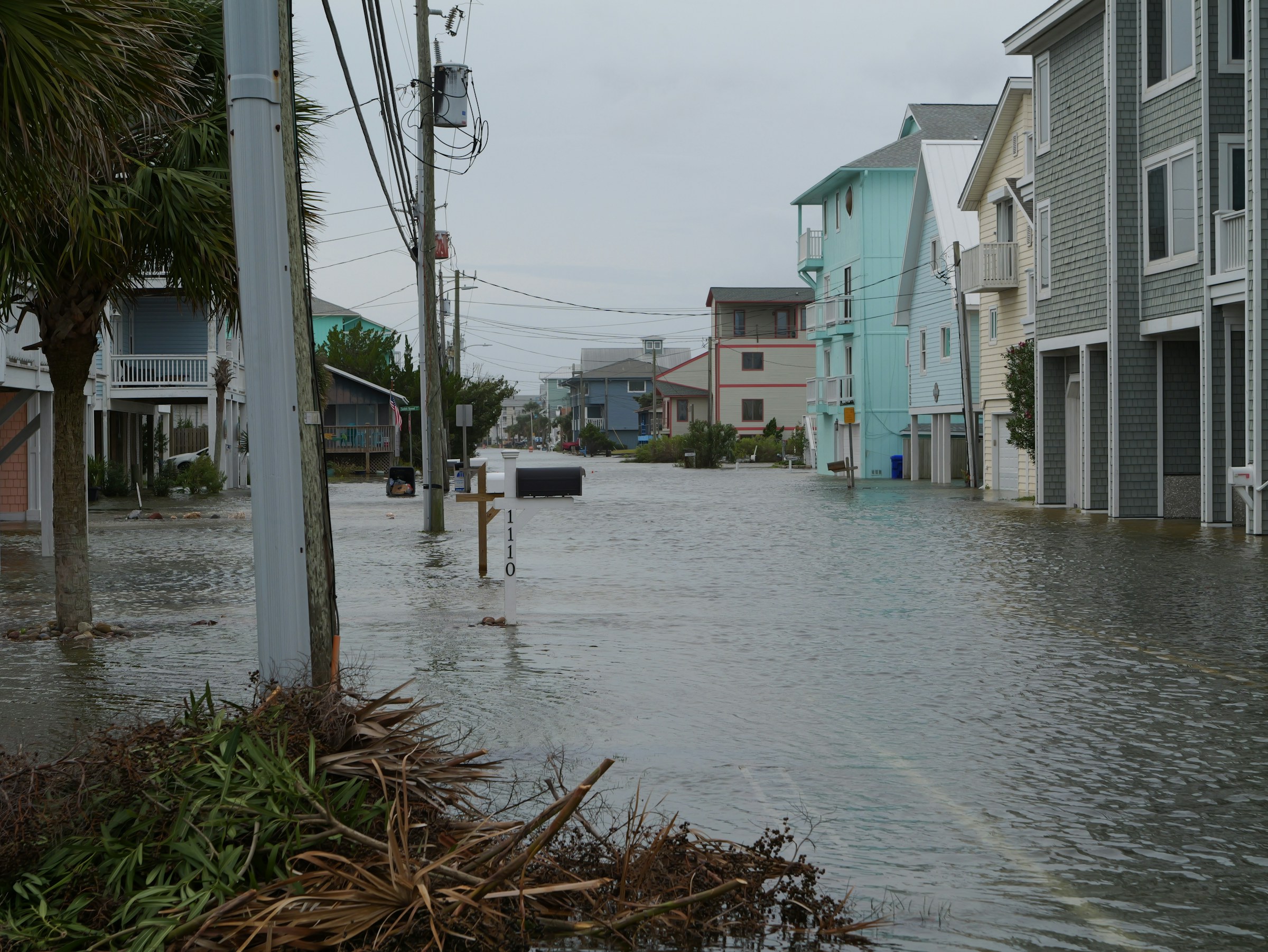 A flooded beach-town street after a storm, water past the mailbox posts and debris at the curb